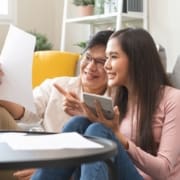 A happy couple sitting on the floor of their living room reviewing financial documents and using a calculator to plan their emergency fund.